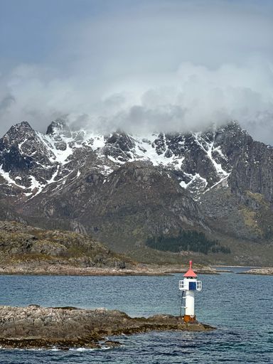 A small lighthouse stands on rocky shores against a backdrop of snow-capped mountains and a cloudy sky in Norway.