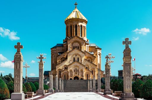 The Holy Trinity Cathedral of Tbilisi, Georgia, under a clear blue sky with ornate crosses and lush greenery.