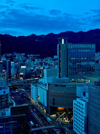 Aerial view of Kobe cityscape at dusk with illuminated modern buildings and mountains in the background.