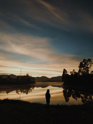 A vertical shot of a person's silhouette on the bay of a lake in a mountainous area at sunset