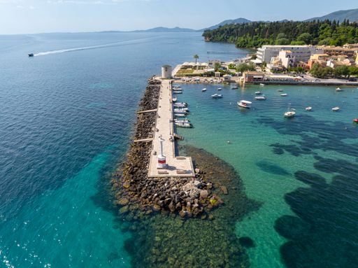 Aerial view of a scenic pier extending into the turquoise sea with boats and a coastal town in the background, under a clear blue sky.