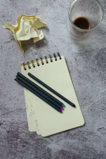 A top view of a spiral notebook with pens, a crumpled paper, and a glass of coffee on a textured surface.