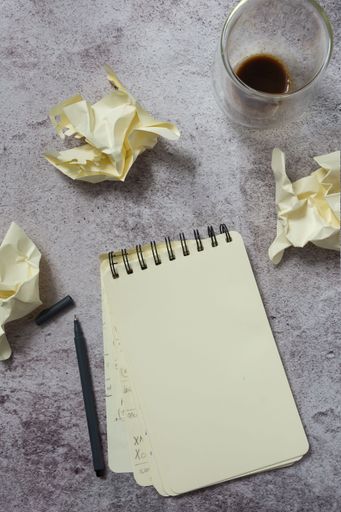 An overhead view of a notepad, pen, crumpled paper, and a half-full coffee cup on a textured surface.