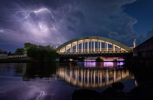 Dramatic lightning over a bridge reflecting in calm waters at night, creating a stunning urban landscape scene.