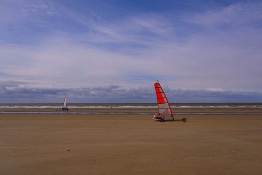 Two land yachts with red sails glide across a vast sandy beach under a partly cloudy sky, capturing the essence of outdoor recreation.