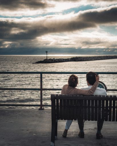 An adult couple sitting on a bench and looking at the sea under a cloudy sky