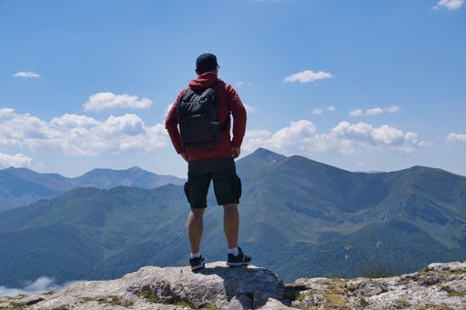 A man in his 30s with a backpack stands on a mountain peak, overlooking a vast landscape