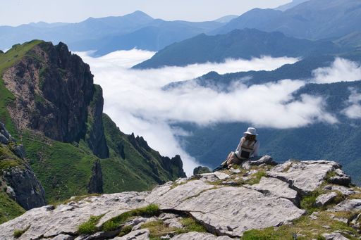 A woman in a sun hat sits on a rocky mountain ledge, overlooking a breathtaking landscape