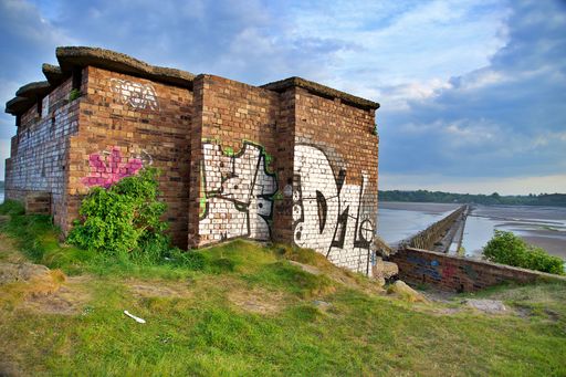 Old brick building with graffiti by a riverbank under a cloudy sky, showcasing urban art and nature.