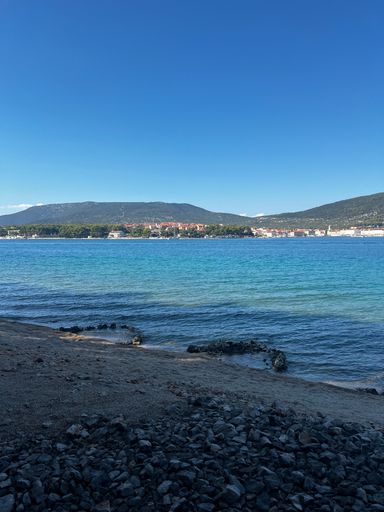 A scenic view of a rocky beach with clear blue water and distant mountains under a bright blue sky in Croatia.