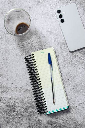 A flat lay of a to-do list notebook with a pen, smartphone, glass of coffee on a textured surface.