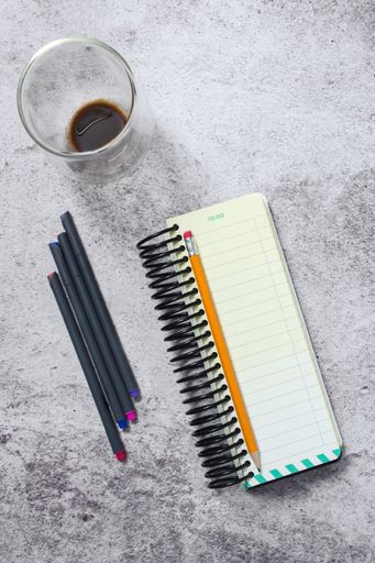 A top view of a to-do list notebook with a pencil, pens, and a glass of coffee on a textured surface.