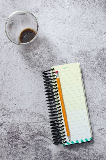 A top view of a to-do list notebook with a pencil and a glass of coffee on a textured surface.