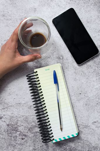 Top view of a hand holding a coffee cup next to a smartphone, spiral notebook, pen on a textured surface.
