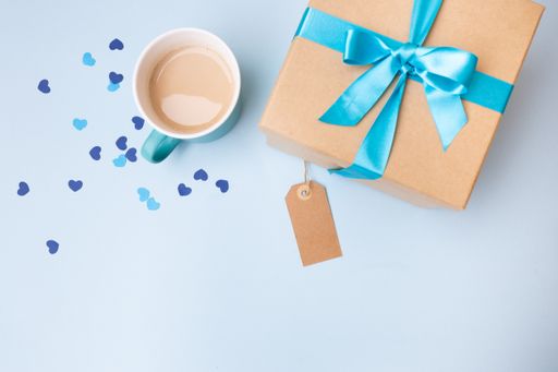 A top view of a gift box wrapped with a blue ribbon on a blue surface with a cup of coffee on it