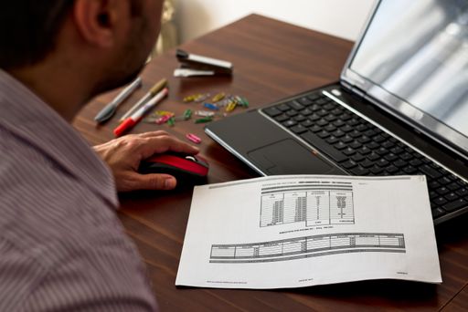 An over-shoulder shot of a male doing some work on his laptop, sitting at the table surrounded by different stationery items around him