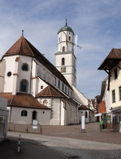 A historic church with a tall clock tower in a quaint European town under a partly cloudy sky.