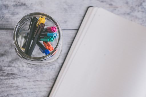 An overhead shot of colorful markers in a jar next to an opened textbook on a wooden surface