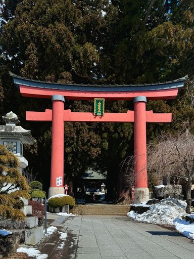 A red torii gate in a Japanese shrine surrounded by trees and snow, showcasing traditional architecture and serene winter scenery.