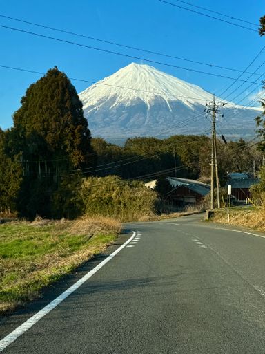 A scenic road leading to Mount Fuji under a clear blue sky, with lush greenery and power lines in the foreground.