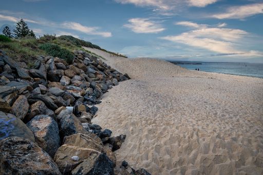 A serene beach scene with sandy dunes and rocky shoreline under a partly cloudy sky, featuring distant figures by the water.