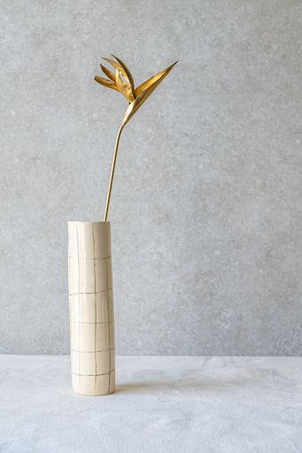 A golden bird of paradise flower stands tall in a light-colored cylindrical vase with a subtle grid pattern.