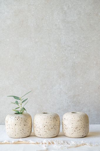 Three small, speckled concrete vases, one holding a sprig of greenery, sit against a textured neutral background.