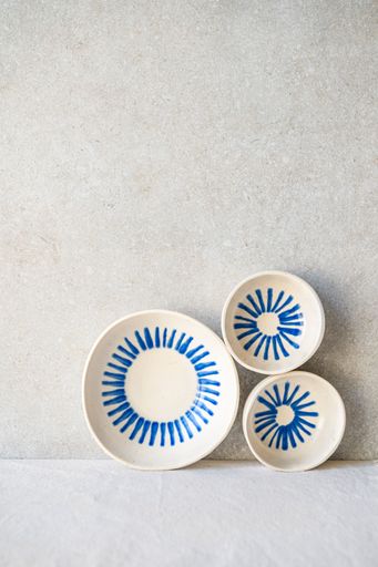 Three white ceramic dishes with a radiating blue hand-painted design are arranged against a neutral backdrop.