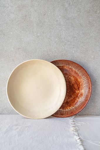 A cream-colored sustainable ceramic dish is placed next to a brown ceramic dish with a textured rim, against a neutral background.