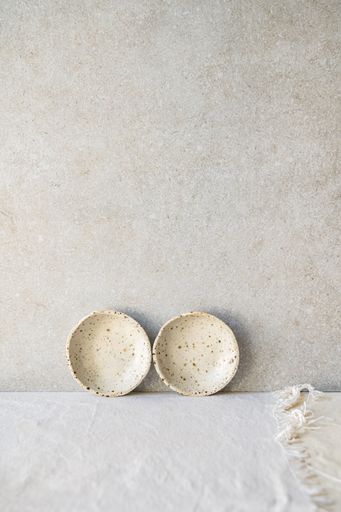 Two small, speckled ceramic dishes are placed against a textured neutral background.