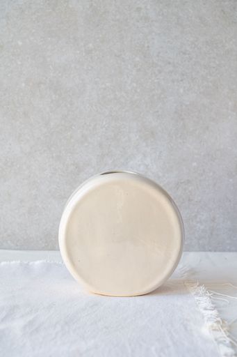A plain, round, off-white ceramic vase sits on a light surface against a neutral background.