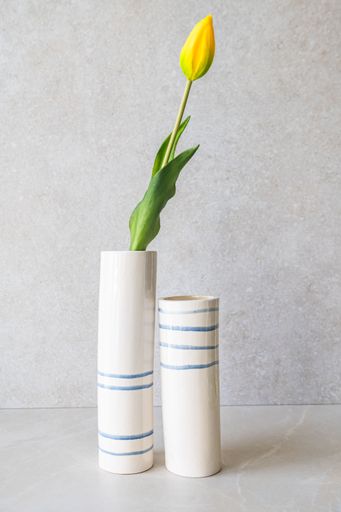 Two white, cylindrical vases, one taller than the other, are decorated with horizontal cobalt blue stripes, with one holding a single yellow tulip.