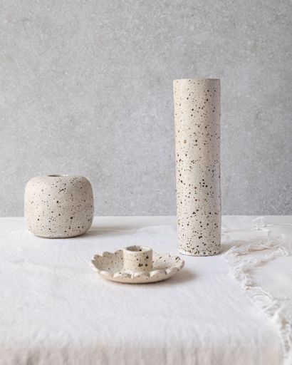 Minimalist speckled ceramic vases and candle holder on a textured surface, showcasing neutral tones and simple design.