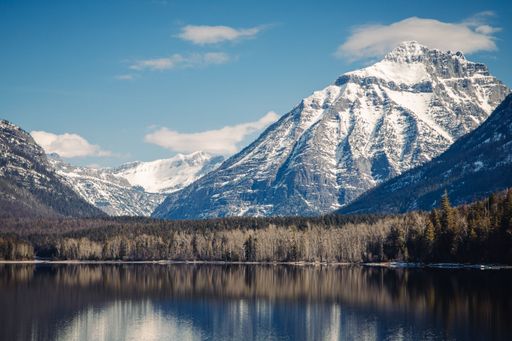 Scenic view of snow-capped mountains reflecting in a tranquil lake under a clear blue sky, surrounded by lush forests.