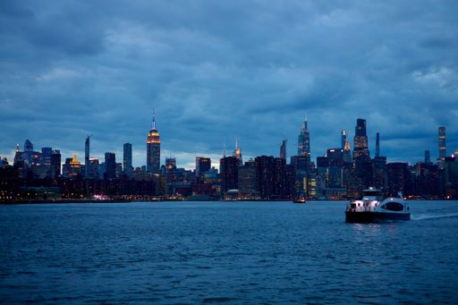 New York City skyline at dusk with a ferry crossing the Hudson River, highlighting iconic skyscrapers under a cloudy sky.