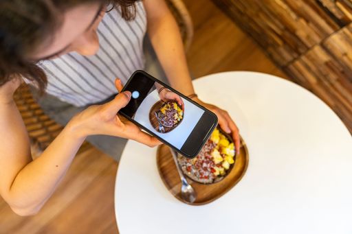 Customer photographing colorful acai bowl with smartphone, capturing vibrant dessert in modern cafe setting.