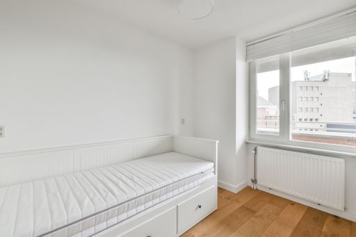 An interior shot of a bedroom in white tones with a white bed next to a window