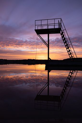 A silhouette of a diving platform reflecting on calm water during a vibrant sunset