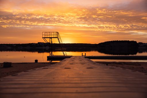 A tranquil lakeside scene at sunset with a diving platform silhouetted against a vibrant sky