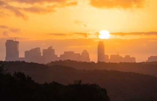 Silhouette of a city skyline at sunset with a vibrant orange sky and sun setting behind buildings.