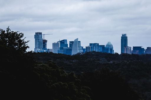 Skyline of Austin, Texas with modern skyscrapers against a cloudy sky, viewed over a lush green landscape.