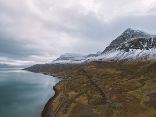 Scenic view of a snow-capped mountain and a winding road along a tranquil lake under a cloudy sky in Iceland.
