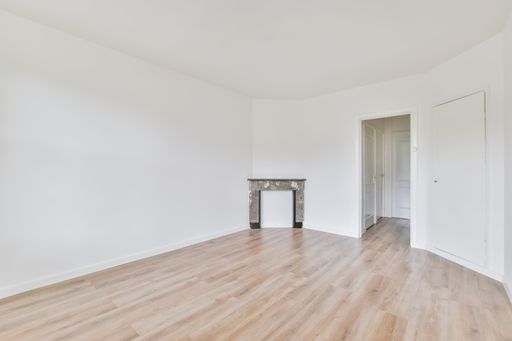An interior shot of an empty room with white walls and a wooden floor with an empty fireplace