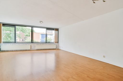 An interior shot of an empty room with white walls and a wooden floor next to a window