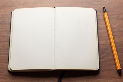 An open blank notebook with a black binding and a black ribbon bookmark lies on a wooden surface next to an orange and black pen.