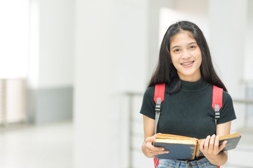 A young cheerful Southeast-Asian female college student carrying book stands in a campus building