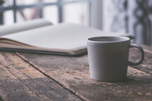 A closeup shot of a white ceramic mug with coffee in it on a wooden surface next to a textbook