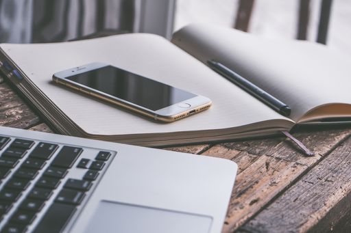 A closeup shot of a smartphone on an opened textbook with a laptop on the side with a wooden background