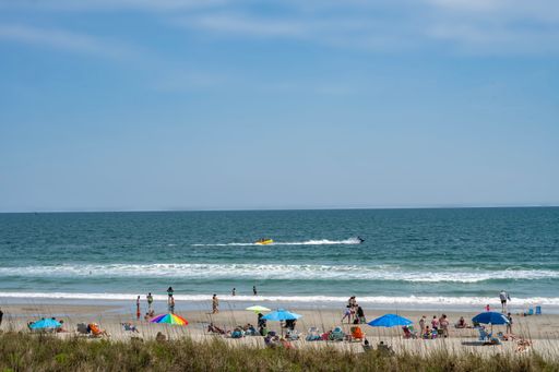 Sunny day at a beach with people relaxing under colorful umbrellas and a jet ski in the ocean.