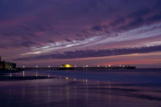 A stunning view of a pier at sunset with vibrant purple and pink skies reflecting on the calm ocean waters.